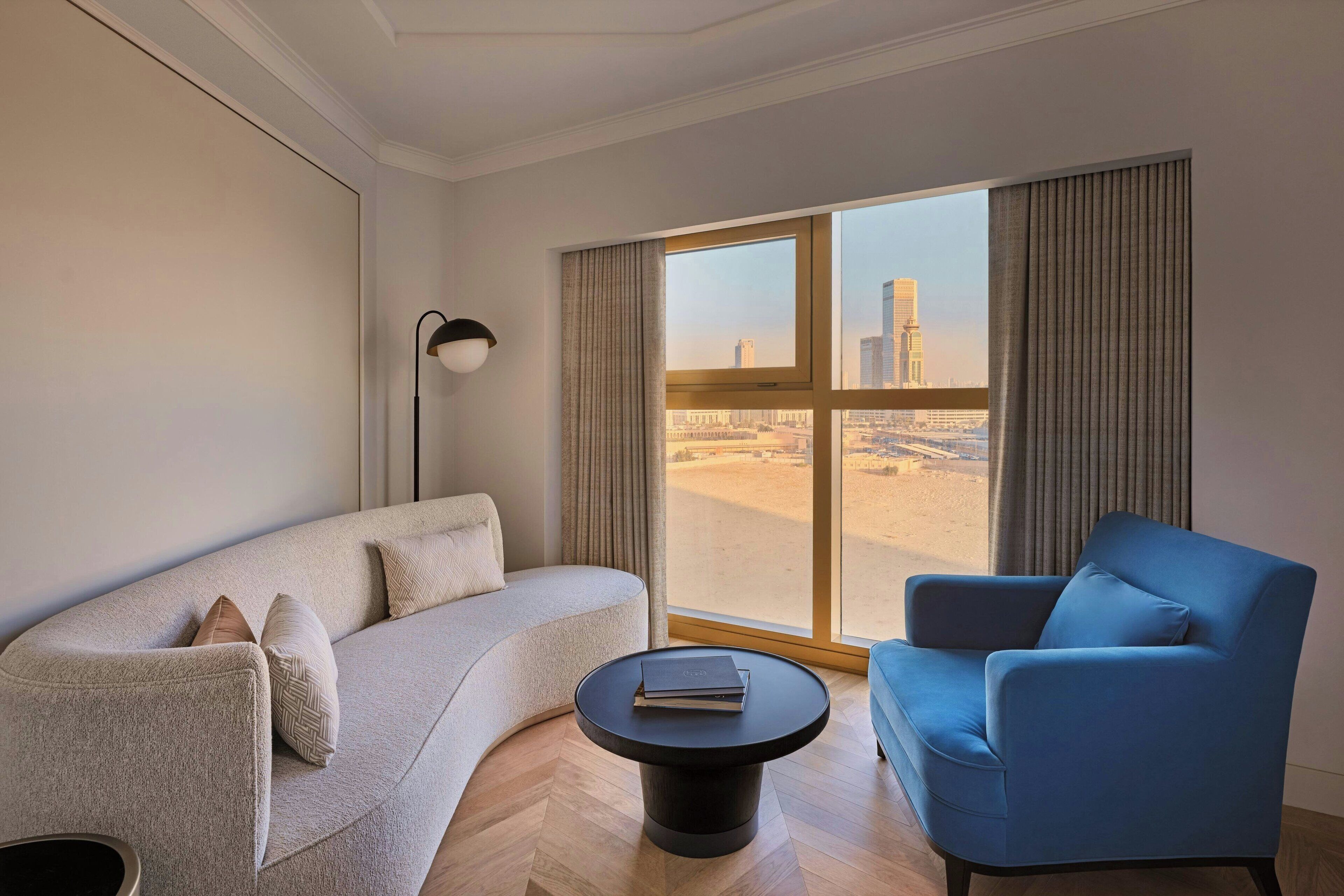Una habitación de hotel moderna con un sofá curvo, un sillón azul, una mesa de centro redonda y un amplio ventanal con vista a la ciudad.