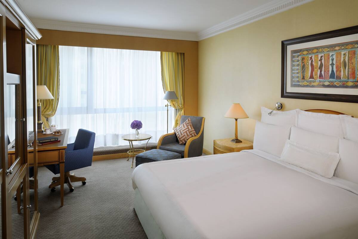 Une chambre d’hôtel avec un grand lit, un bureau avec une chaise, une petite table avec une lampe et une fenêtre avec des rideaux.