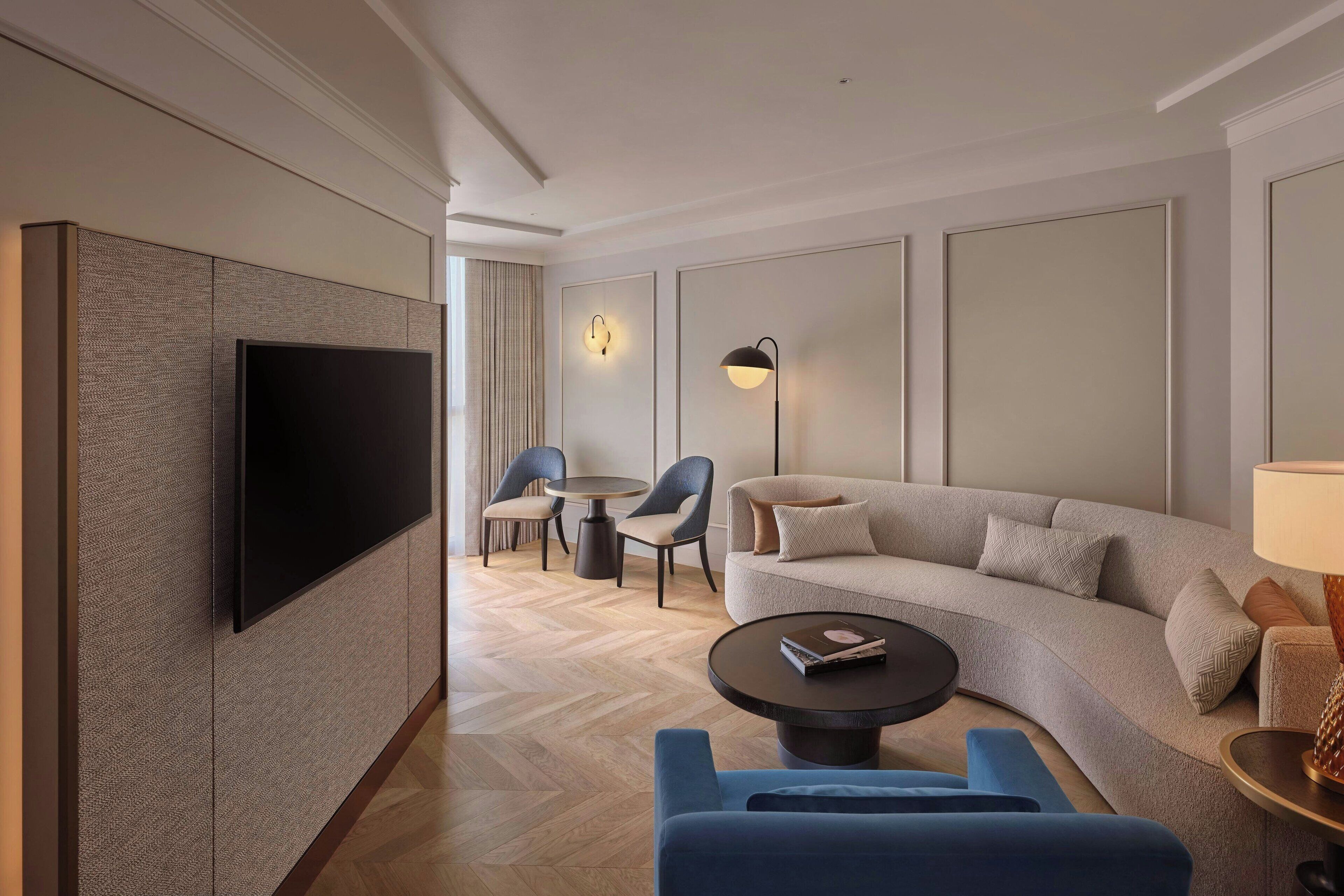 Una habitación de hotel moderna con un televisor de pantalla plana grande montado en la pared, un sofá gris, un sillón azul, una mesa de centro redonda y una lámpara de pie.