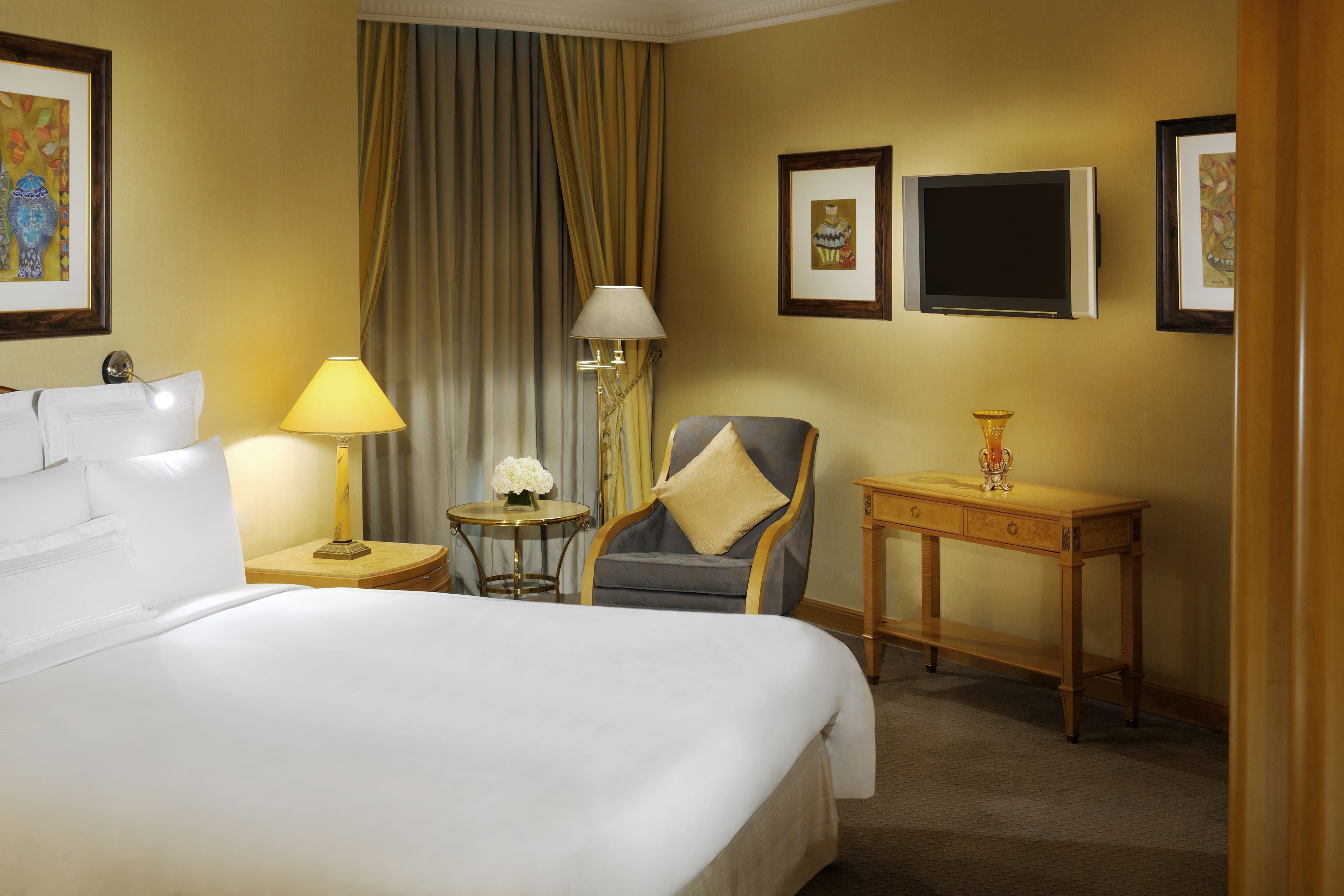 A hotel room with a large bed, a chair, a small table with a lamp, and a TV on the wall.