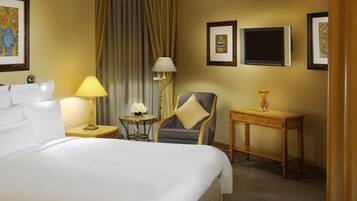 A hotel room with a large bed, a chair, a small table with a lamp, and a TV on the wall.