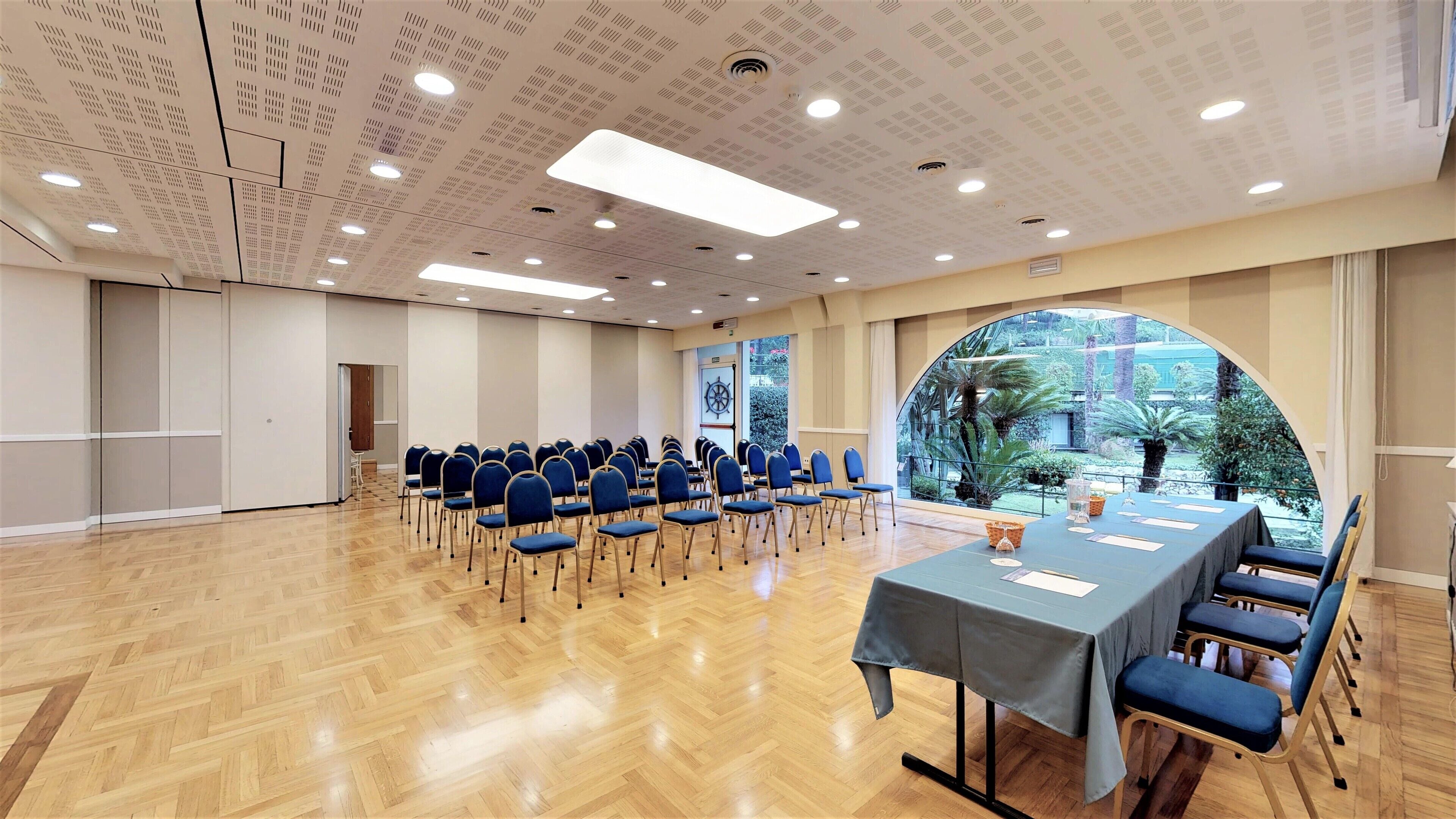Une salle de conférence avec une longue table, des chaises bleues et une vue sur un jardin, visible à travers une grande fenêtre en arc de cercle.