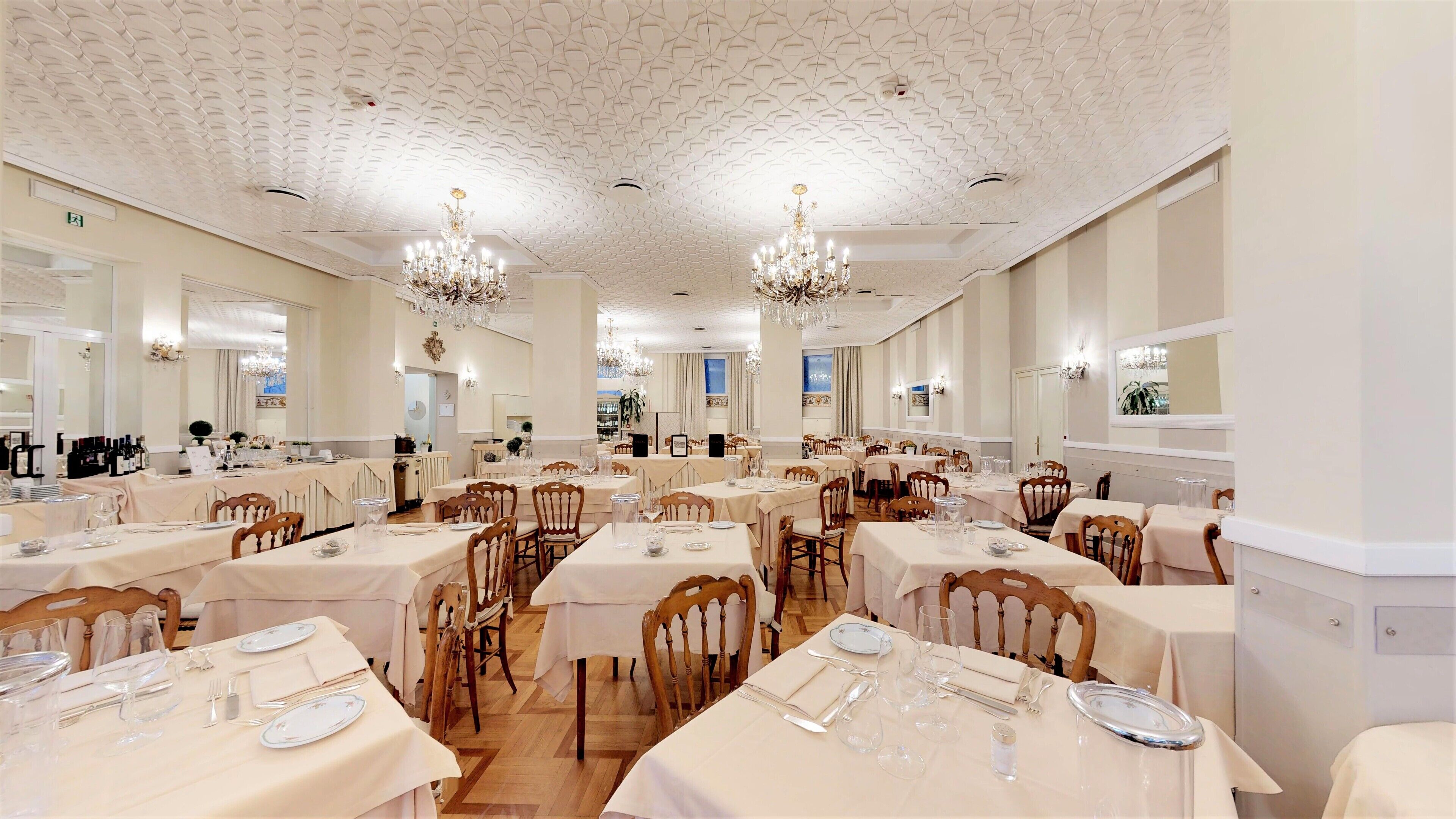 Un comedor amplio con varias mesas y sillas, elegantes candelabros y un techo alto con un diseño decorativo.