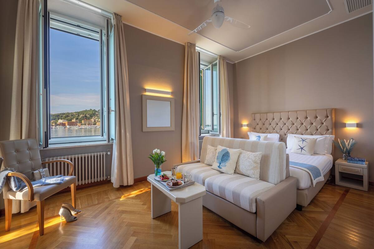 Une chambre d’hôtel avec une grande fenêtre, un canapé, un fauteuil, une petite table sur laquelle se trouve un plateau de nourriture et un lit avec une tête de lit.