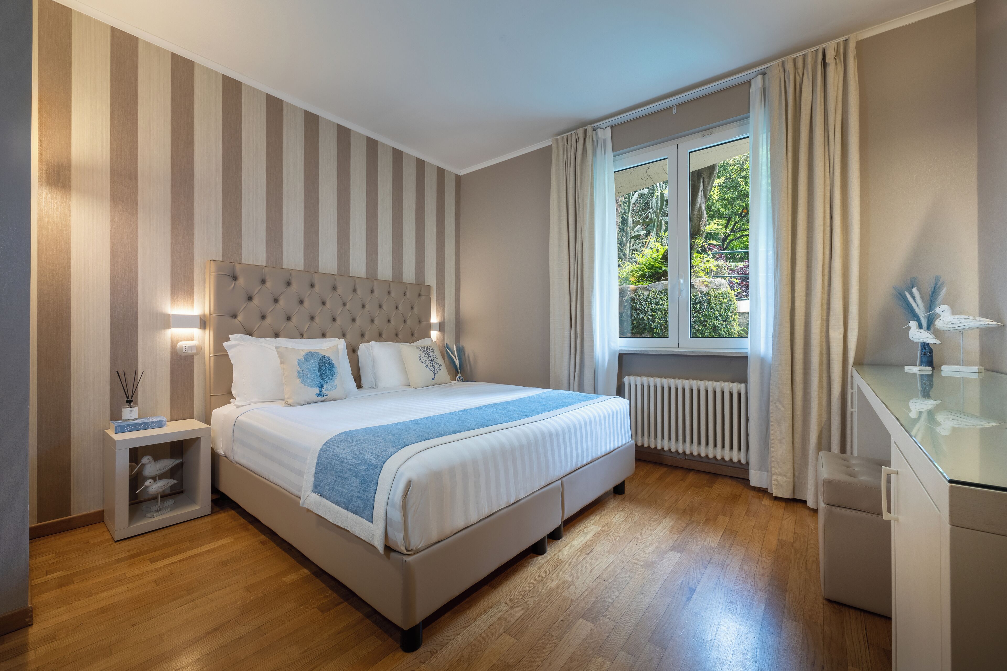 A modern bedroom with a large bed, a bedside table with a lamp, a window with curtains, and a view of greenery outside.