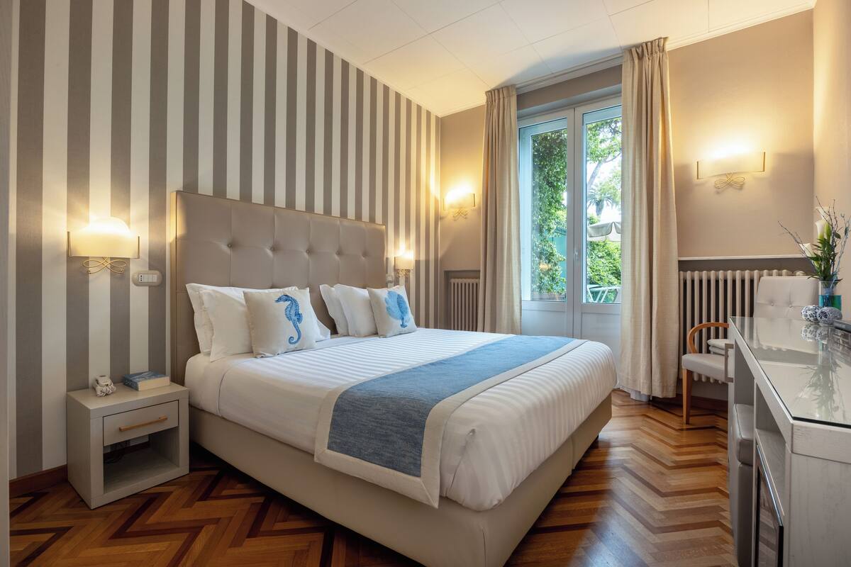 Une chambre d’hôtel avec un grand lit, un papier peint à rayures, une table de chevet, un bureau et une fenêtre avec des rideaux.