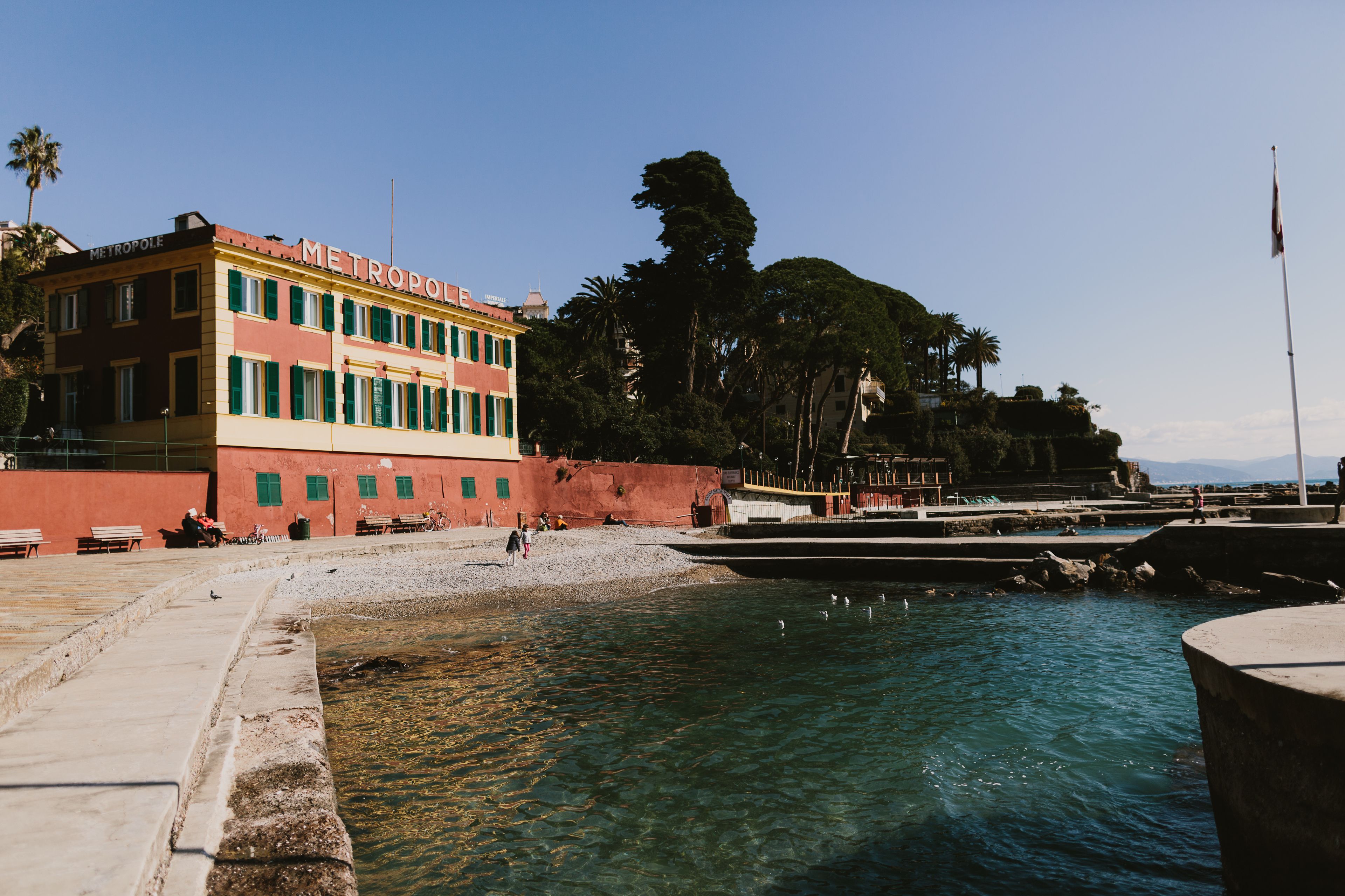 A waterfront hotel with a clear view of the water, a promenade, and a palm tree-lined promenade.