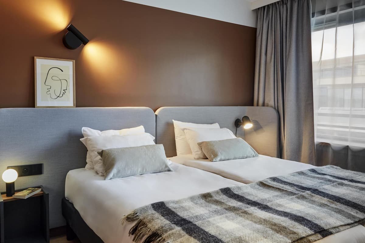 A hotel room with two beds, a bedside table with a lamp, a framed artwork, and a window with curtains.