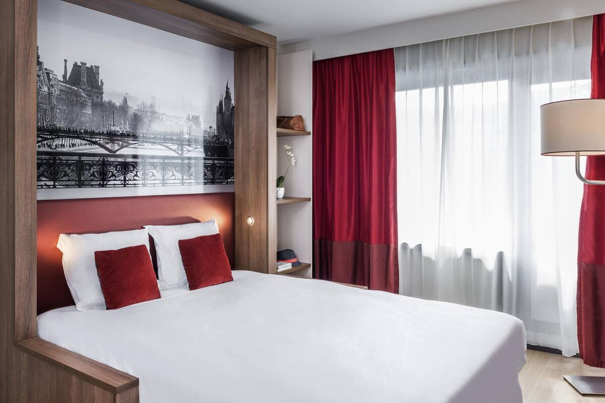 Een hotelkamer met een groot bed, rode gordijnen, een raam met dunne witte gordijnen, een wandlamp en een stadsgezicht schilderij boven het bed.