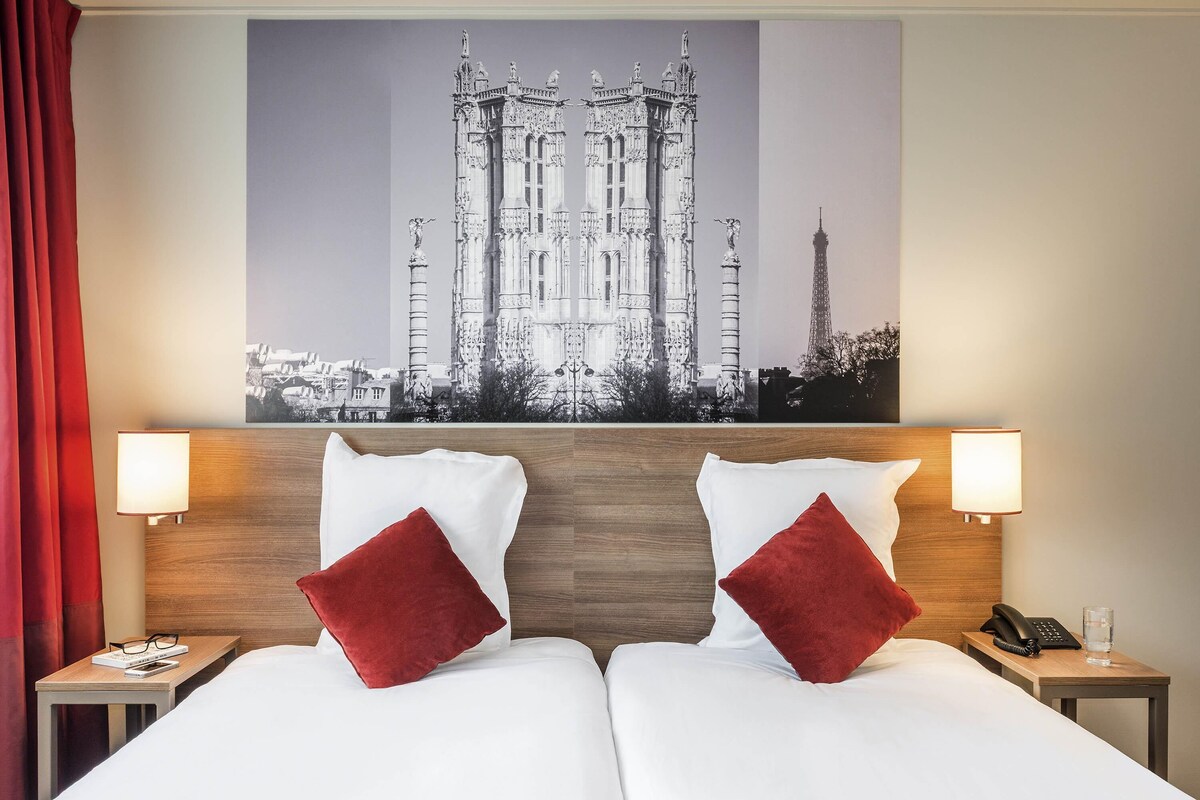 A hotel room with two beds, a wooden headboard, red pillows, bedside lamps, a nightstand with a phone and glass, and a large cityscape painting above the beds.