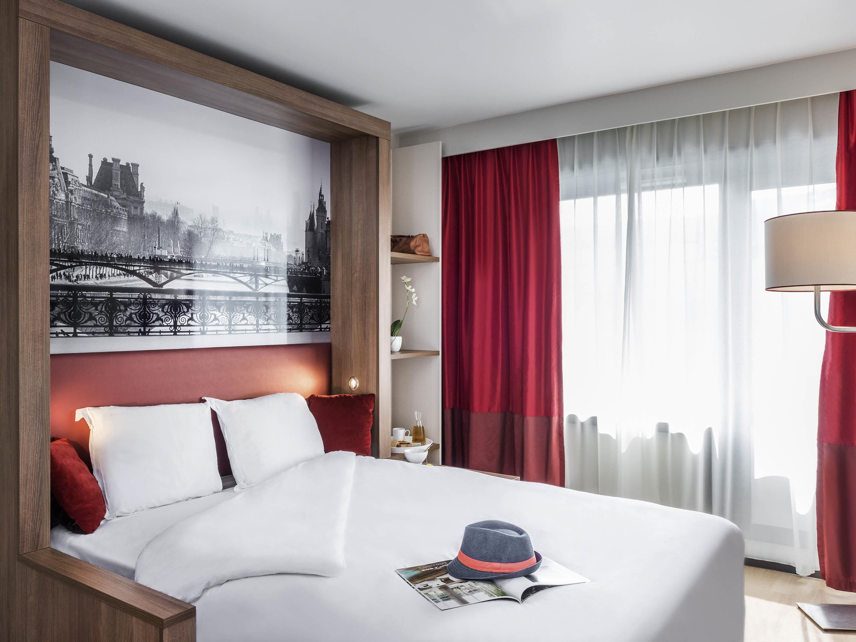 Une chambre d’hôtel avec un lit, une grande œuvre d’art représentant un paysage urbain, un rideau rouge, une lampe et un magazine posé sur le lit.