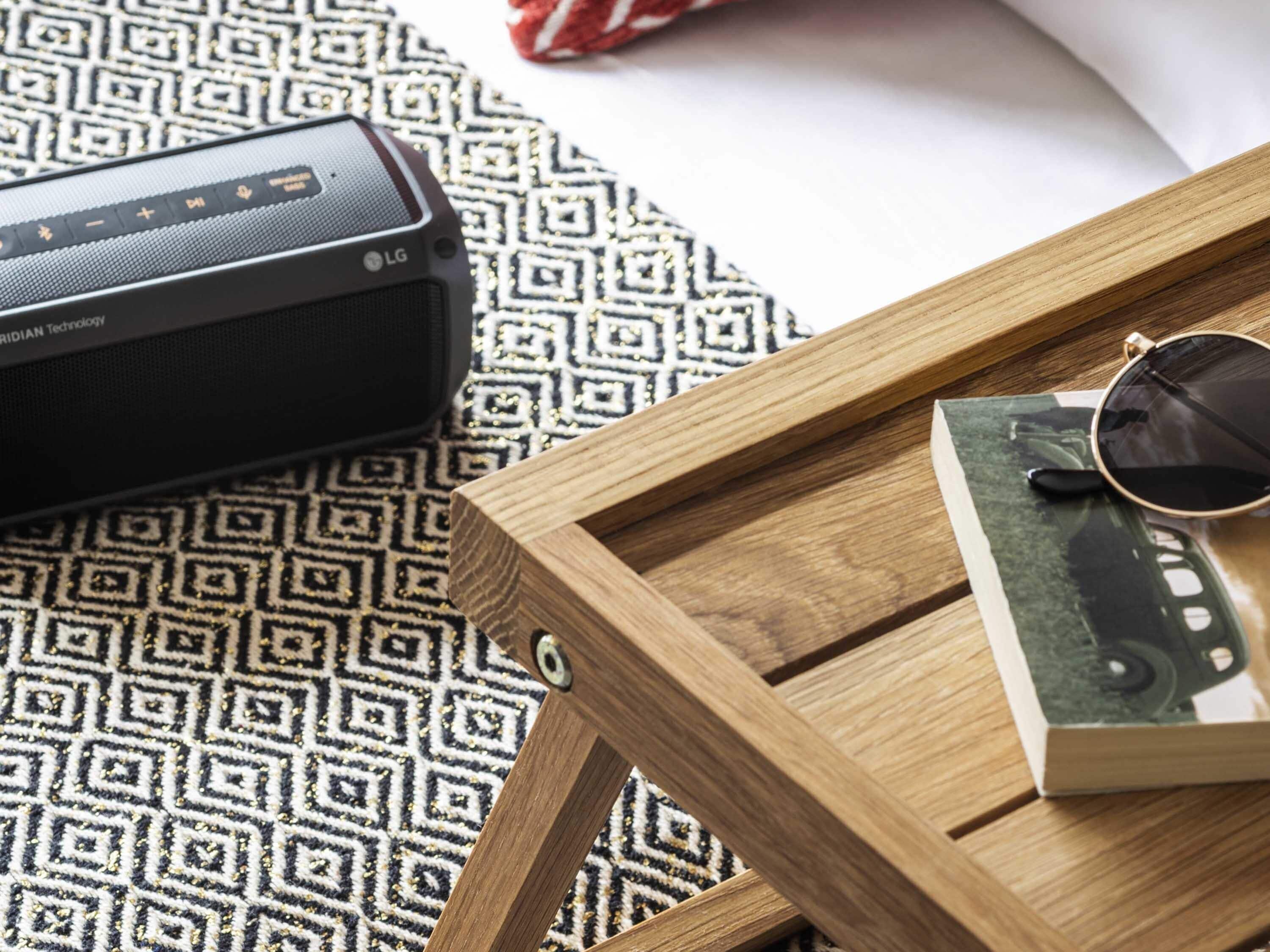 Een zwarte LG-speaker, een zonnebril en een boek op een houten dienblad.