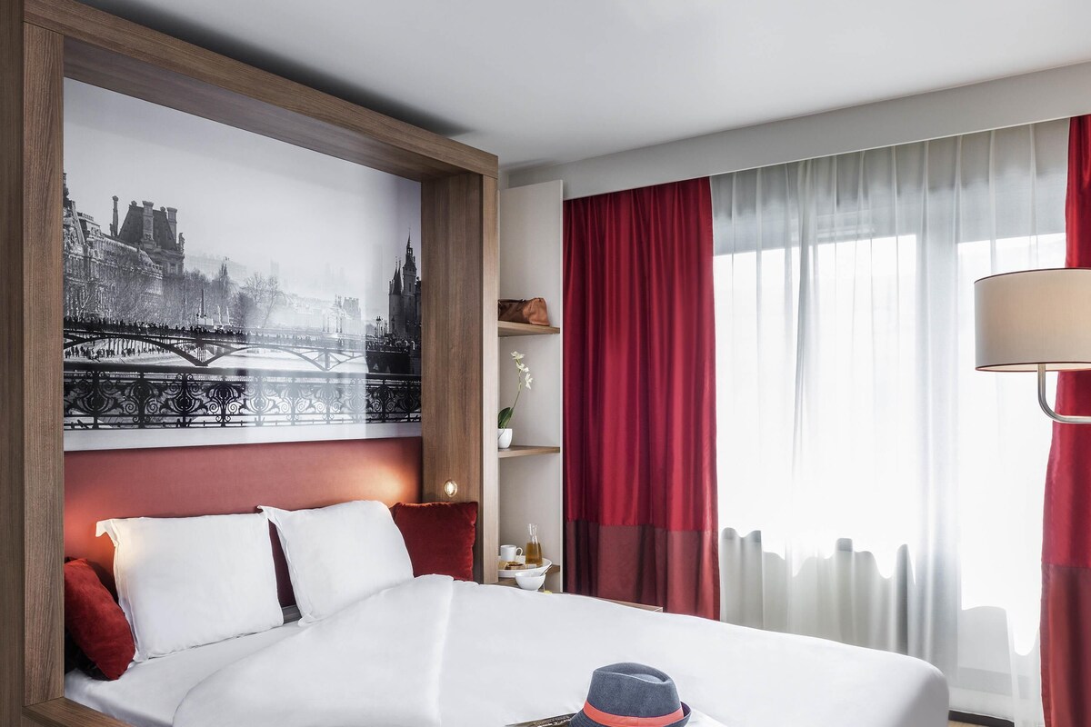 Une chambre d’hôtel avec un lit, une grande œuvre d’art représentant un paysage urbain, un rideau rouge, une lampe et un magazine posé sur le lit.