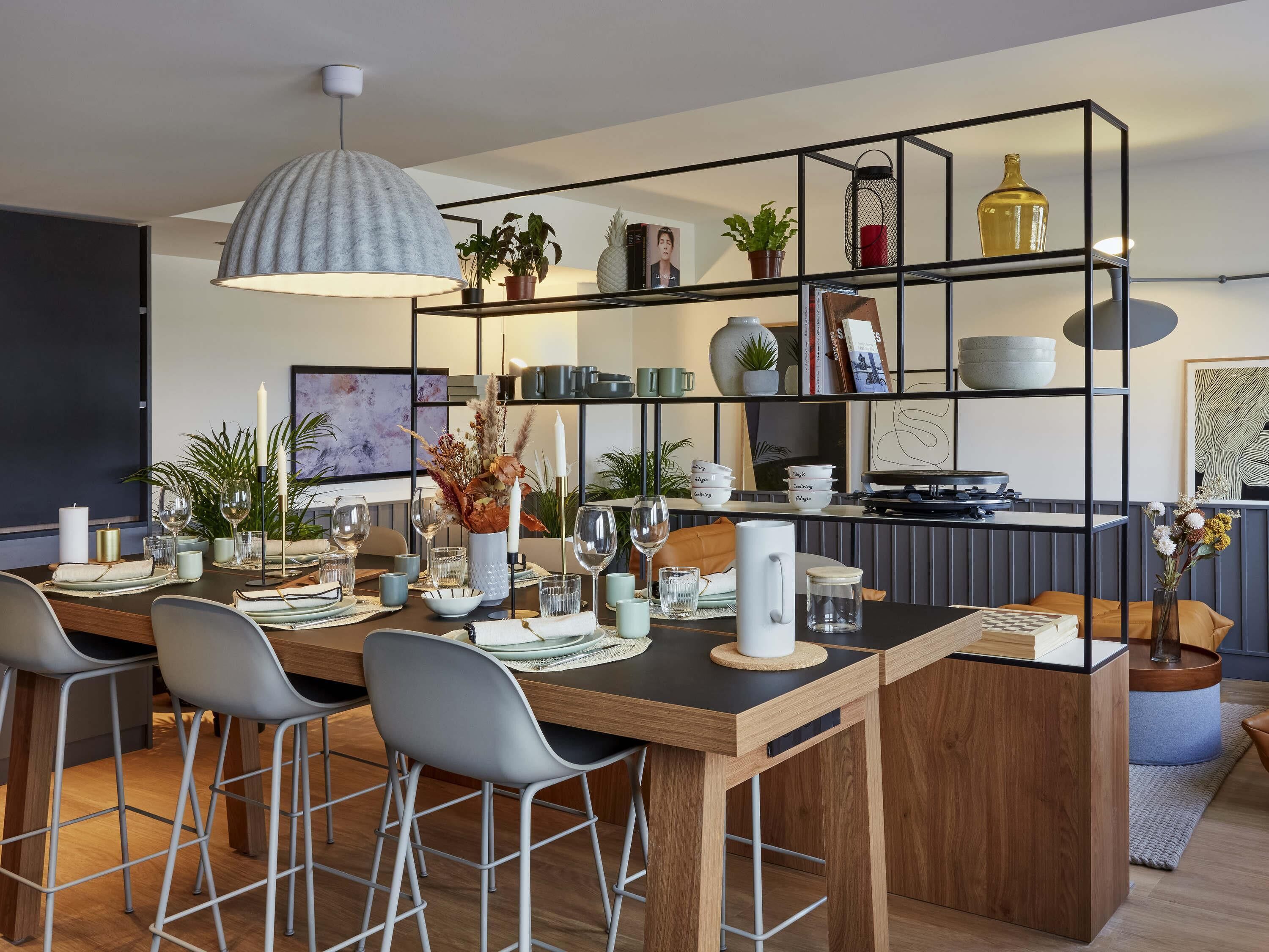 Een moderne keuken met een houten eettafel, barkrukken, een hanglamp en een metalen rek met diverse spullen.