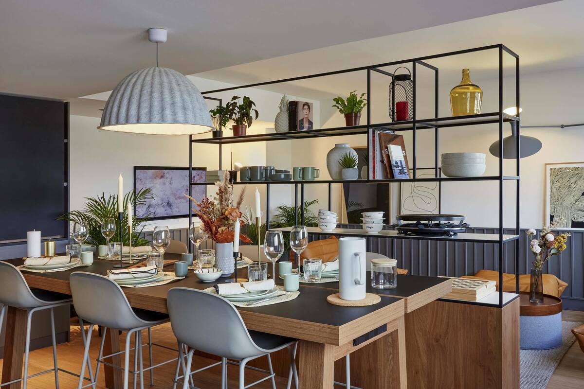 Een moderne keuken met een houten eettafel, barkrukken, een hanglamp en een metalen rek met diverse spullen.