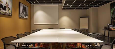 Un salón de conferencias con una larga mesa, sillas de malla negra, un proyector y una pantalla blanca.