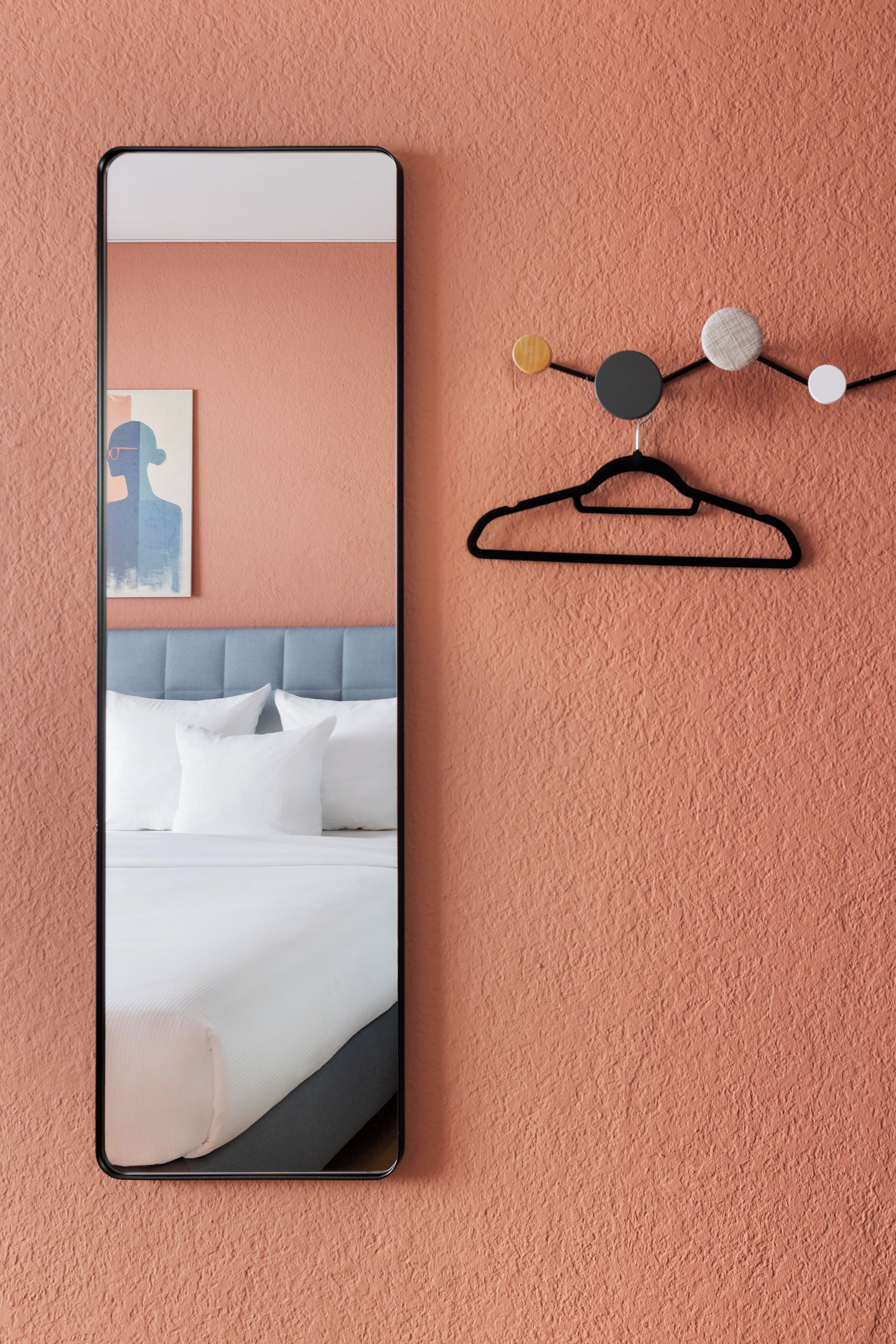 Camera d'albergo con un grande specchio, un letto con lenzuola bianche, una testiera blu e un appendiabiti a parete con motivo circolare.