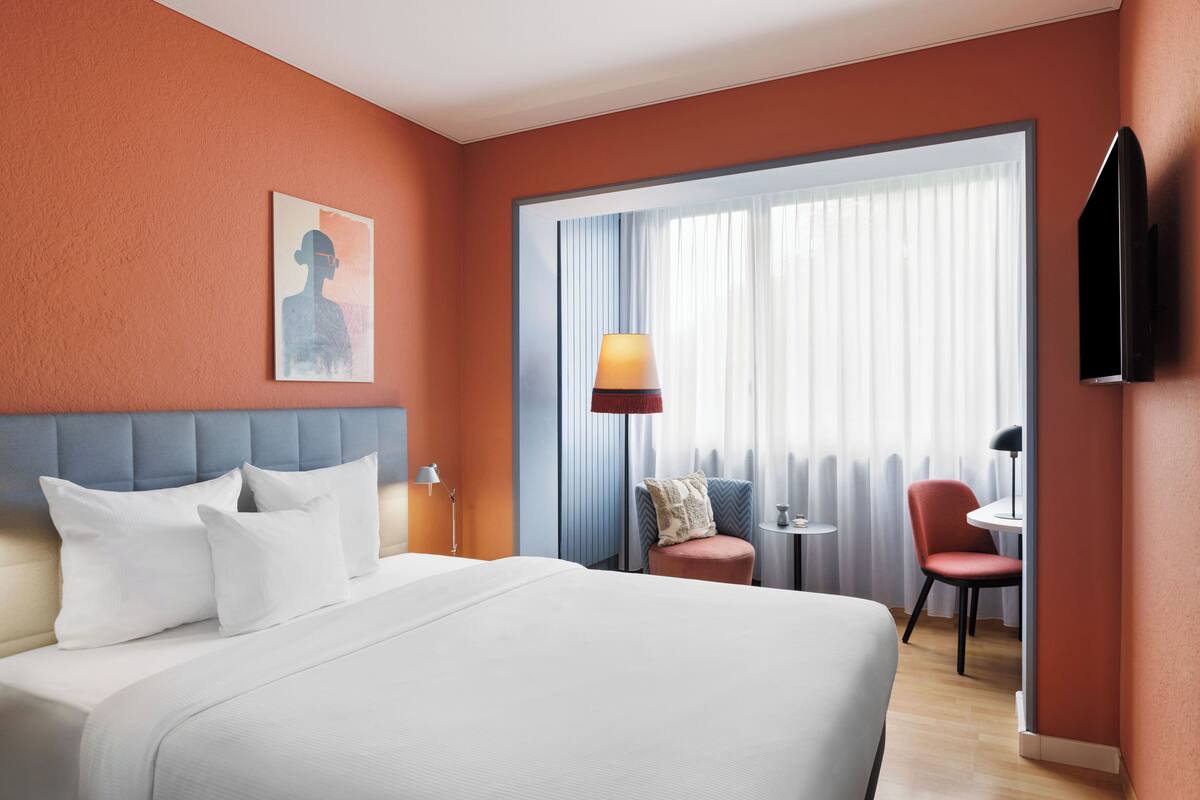Habitación de hotel con una cama grande, una mesita con una silla, un televisor montado en la pared y una ventana con cortinas.
