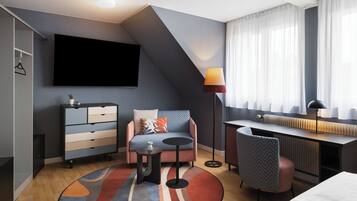 A modern hotel room with a sofa, desk, and chair, a flat-screen TV, and a large window with curtains.