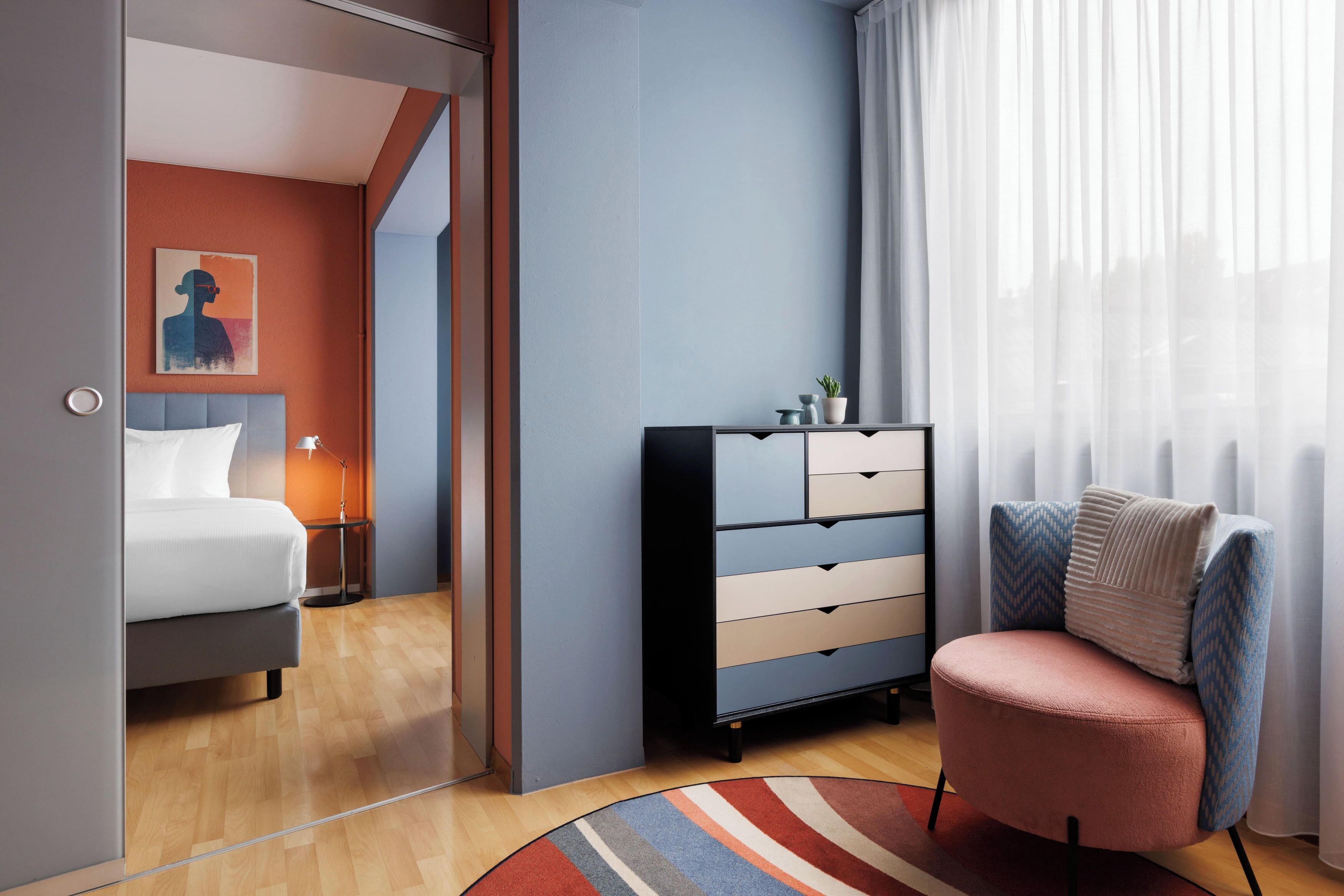 A modern bedroom with a bed, bedside table, dresser, chair, and a view of the outside through a window.