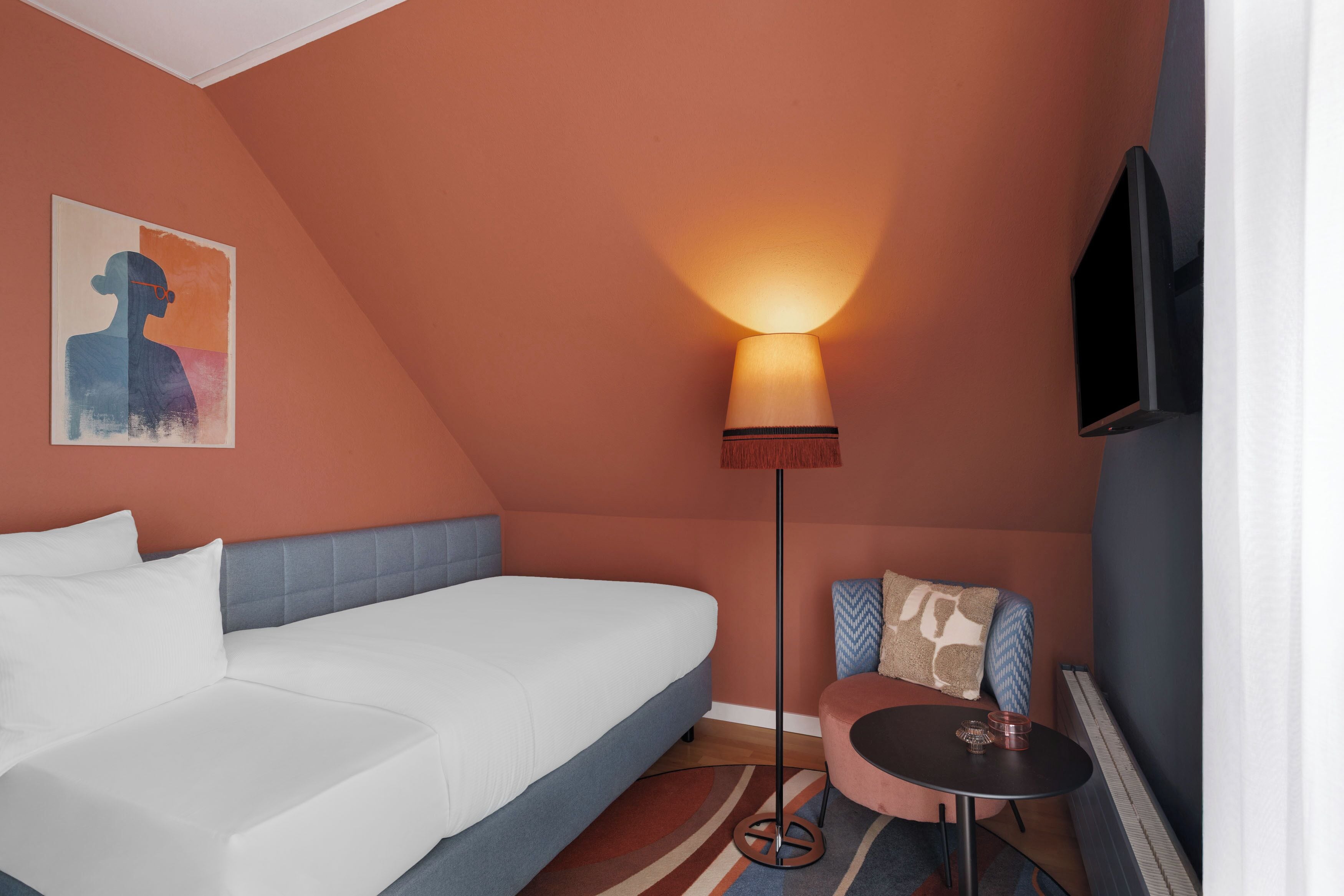 Una habitación de hotel con cama, una mesita, una silla, una lámpara de pie y un cuadro en la pared.