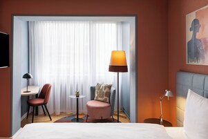 Habitación de hotel con cama, escritorio, silla, una mesita con una planta, lámpara de pie y un cuadro en la pared.