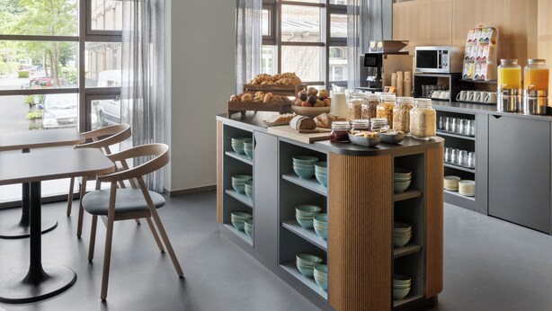 A modern kitchen with a breakfast bar, wooden chairs, and a variety of breakfast items. - Hotel La Pergola (Bern)