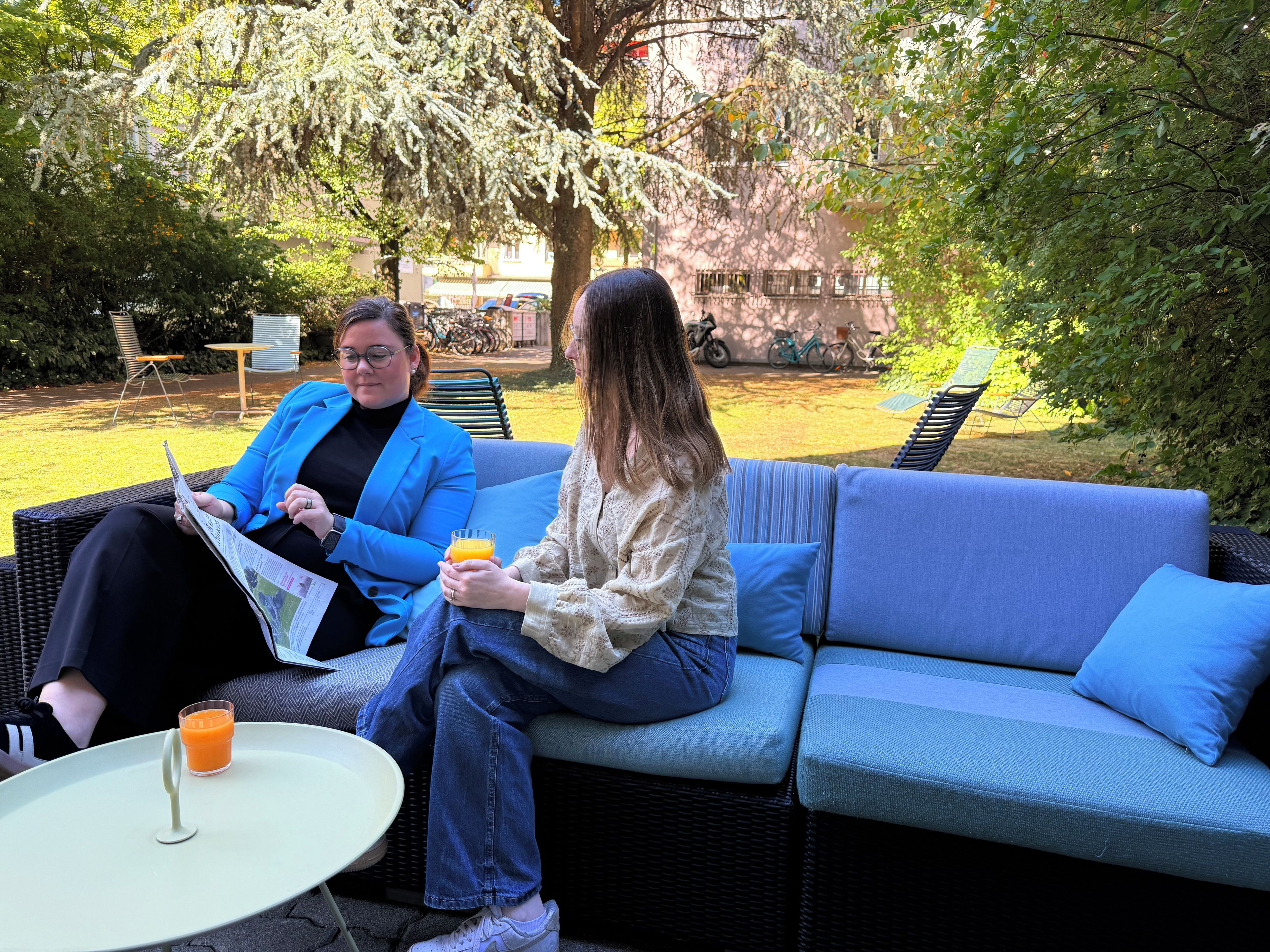Deux personnes sont assises sur un canapé bleu installé à l’extérieur, devant une petite table et un verre de jus d’orange.