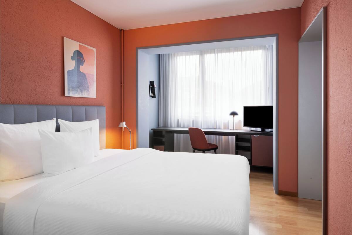 Una habitación de hotel con cama, escritorio y silla, televisor y ventana con cortinas.