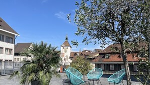 Un área de estar al aire libre con sillas y una palmera, un edificio con una torre, y un cielo azul despejado.