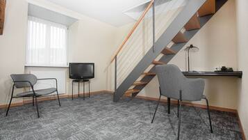 Una habitación con alfombra gris, una silla gris, una silla negra, un televisor, un escritorio con lámpara y una escalera con escalones de madera y barandillas de metal.