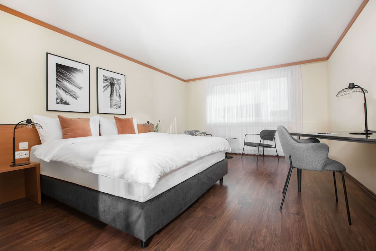 Habitación de hotel moderna con una cama grande, un escritorio y una silla. Hay dos cuadros en la pared, una lámpara en el escritorio y una ventana con cortinas translúcidas.