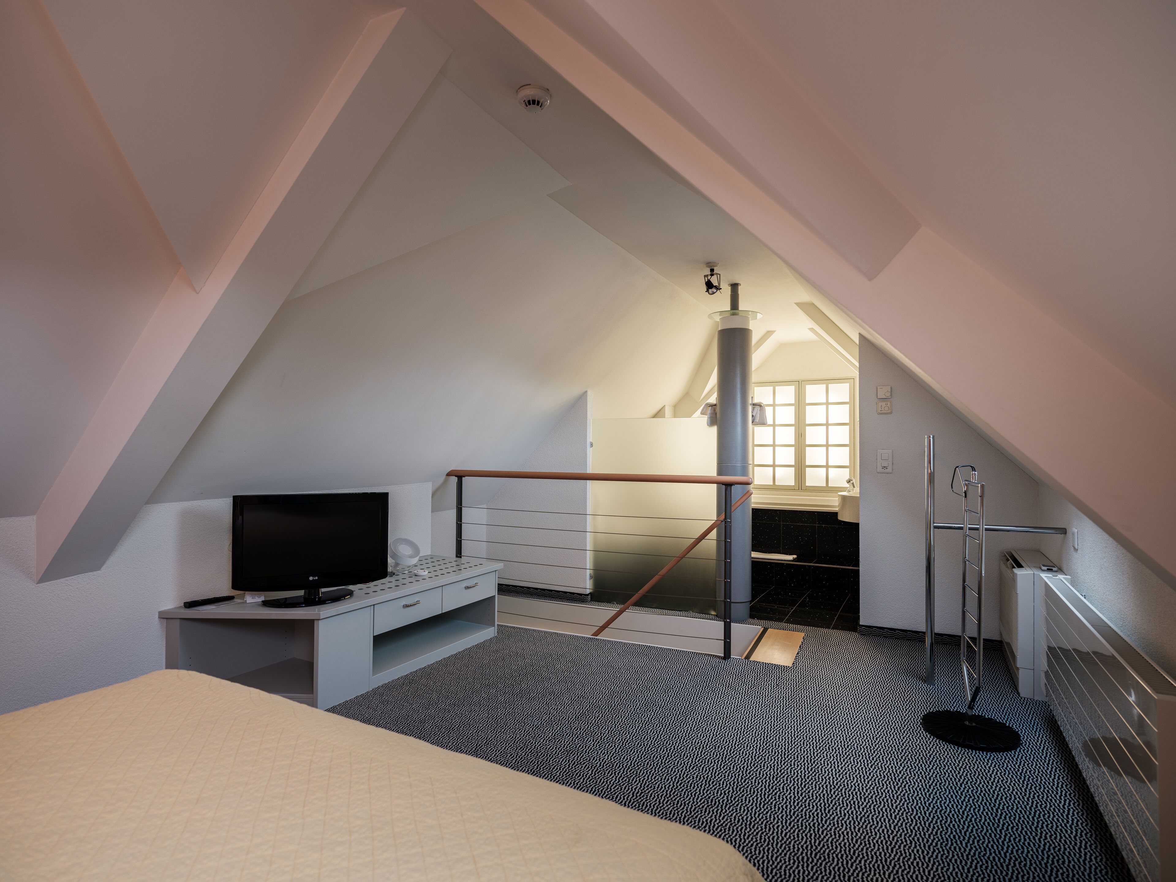 A bedroom with a sloped ceiling, a bed, a desk with a TV, a window, and a bathroom visible in the background.