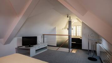 A bedroom with a sloped ceiling, a bed, a desk with a TV, a window, and a bathroom visible in the background.