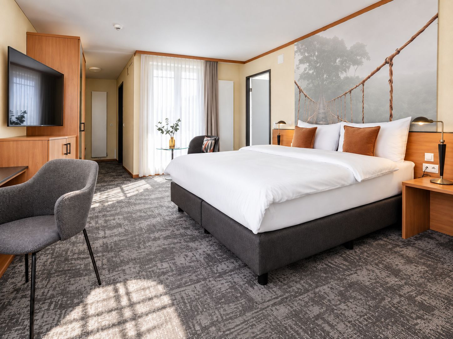 A hotel room with a large bed, a desk, a TV, and a view of a tree through a rope bridge.