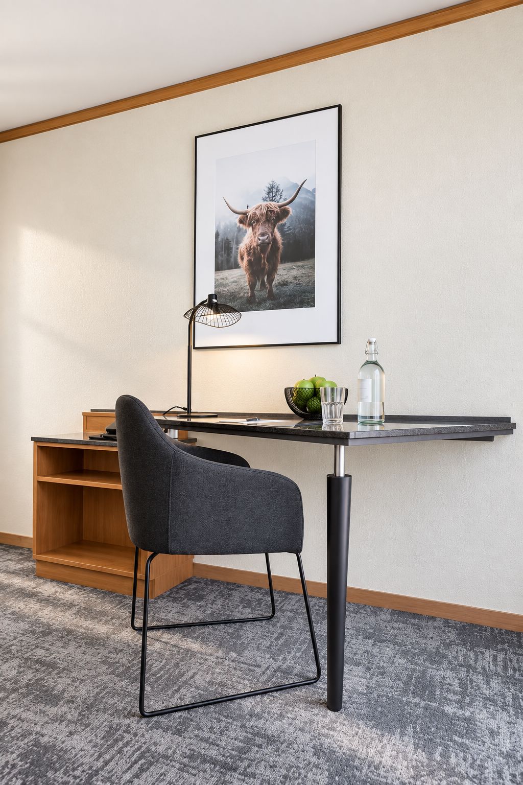 A hotel room with a desk, a framed picture of a Highland cow, a chair, and a small table with a bottle and apples.