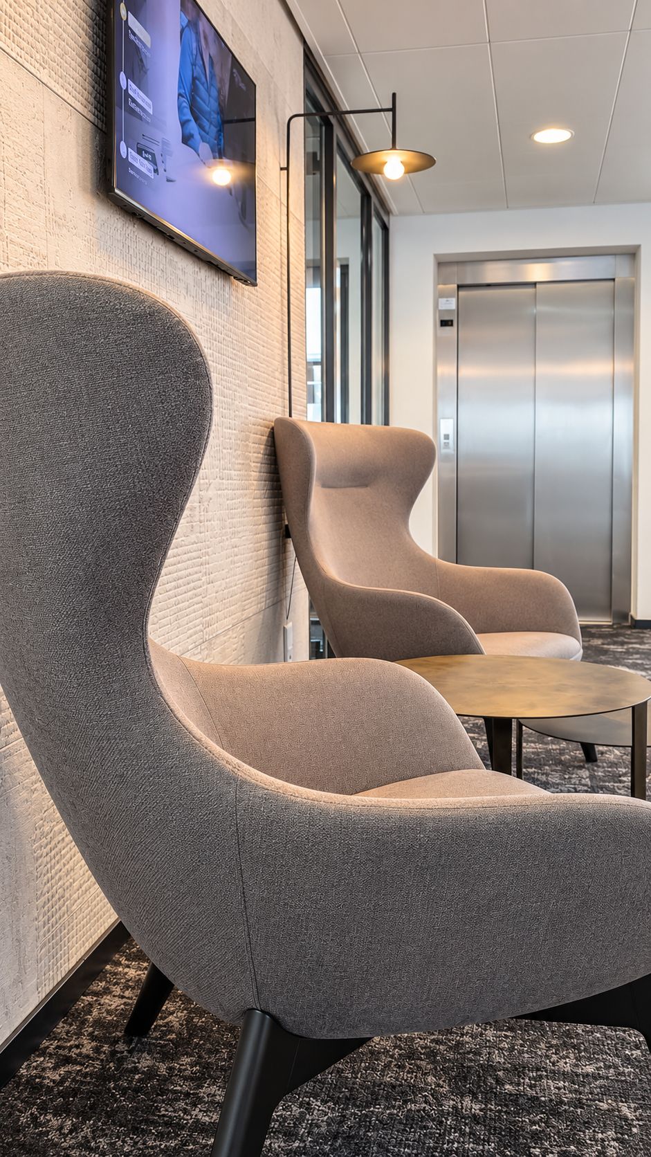 A modern waiting area with two upholstered chairs, a small table, a mounted screen, and an elevator in the background.