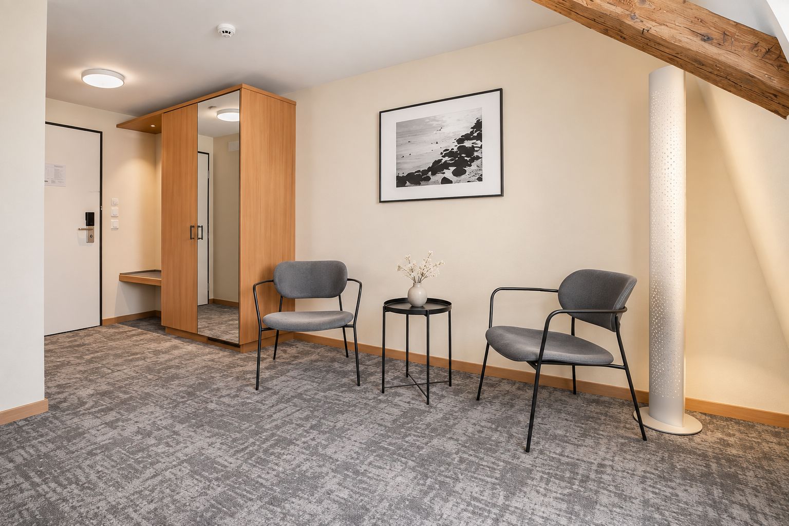 Ein Hotelzimmer mit sichtbarem Holzbalken an der Decke, einem gerahmten Bild an der Wand, zwei grauen Stühlen, einem kleinen Tisch mit einer Vase und einem Teppichboden.