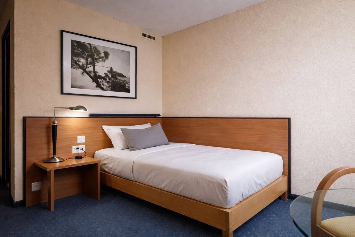 Habitación de hotel con cama de madera, mesita de noche con lámpara, toma de corriente e imagen enmarcada en la pared.