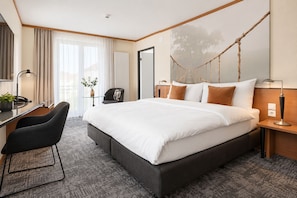 Een moderne hotelkamer met een groot bed, een bureau met stoel, een klein tafeltje met een plant en een hangmat aan de muur.