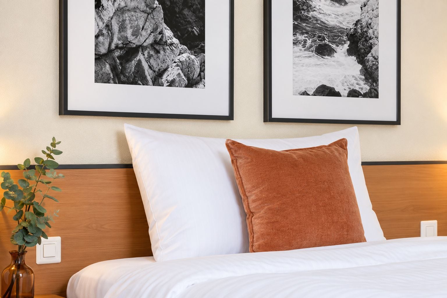 Une chambre d’hôtel avec un lit, deux tableaux encadrés au mur, une plante sur la table de chevet et une tête de lit en bois.