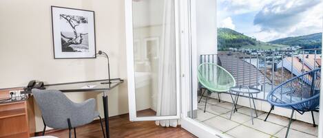 A room with a bed, a desk with a chair, a balcony with chairs, and a view of the landscape.