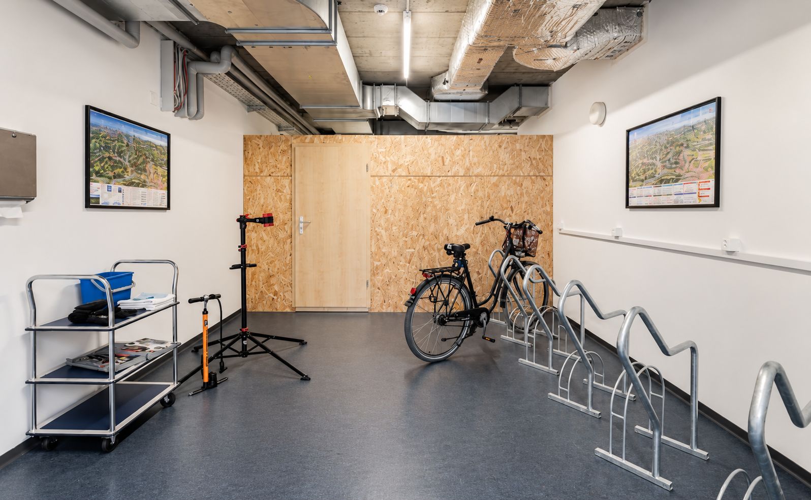 Una habitación con una puerta de madera, una bicicleta, un carro metálico con objetos y un soporte para bicicletas en la pared.