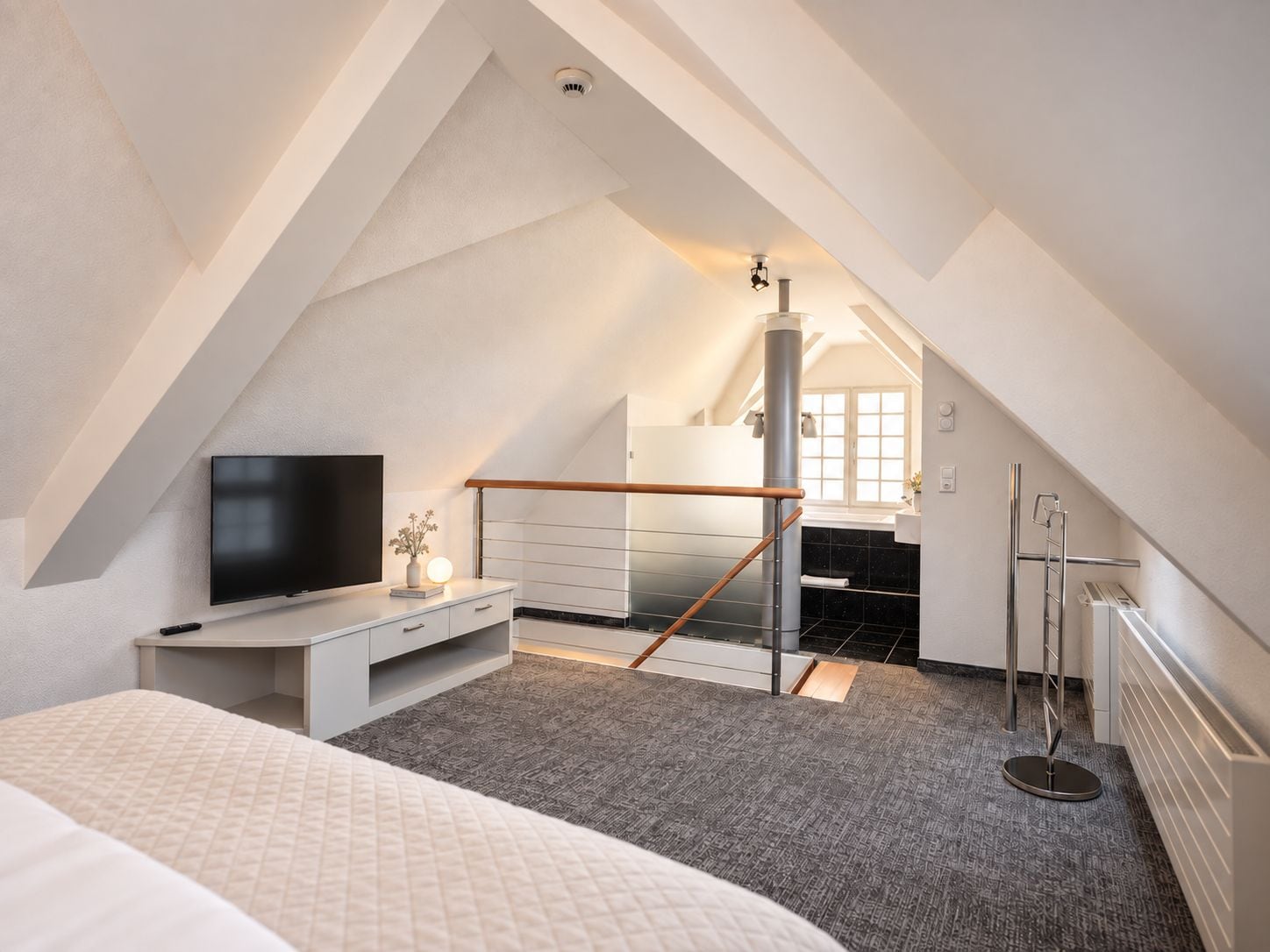 A bedroom with a sloped ceiling, a bed, a desk with a flat-screen TV, a radiator, and a staircase leading to an upper level.