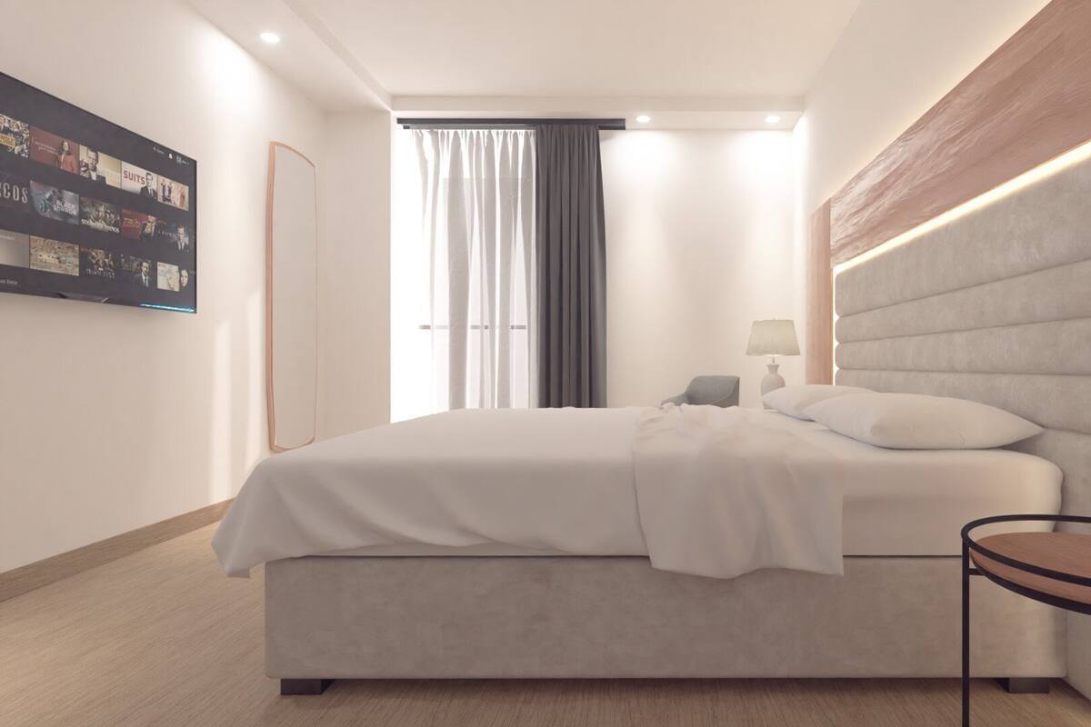 Una habitación de hotel moderna con una cama grande, una mesita de noche, un televisor instalado en la pared y una ventana con cortinas.