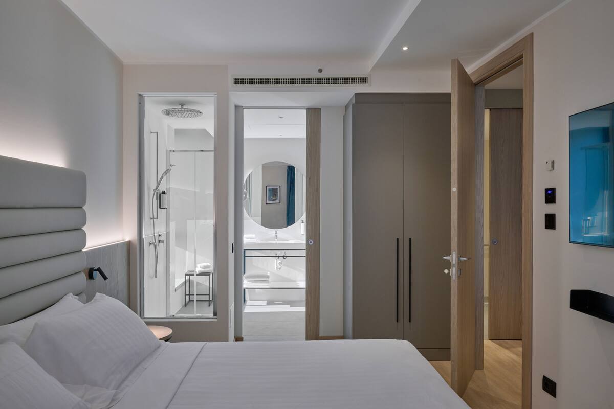 Una habitación de hotel moderna con una cama grande, un vestidor, un baño con ducha y un televisor montado.