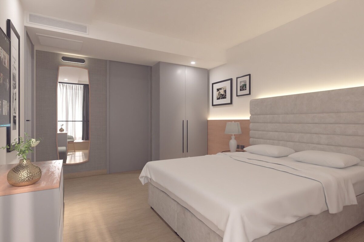 A modern hotel room with a large bed, bedside tables, a desk with a lamp, and a wardrobe.