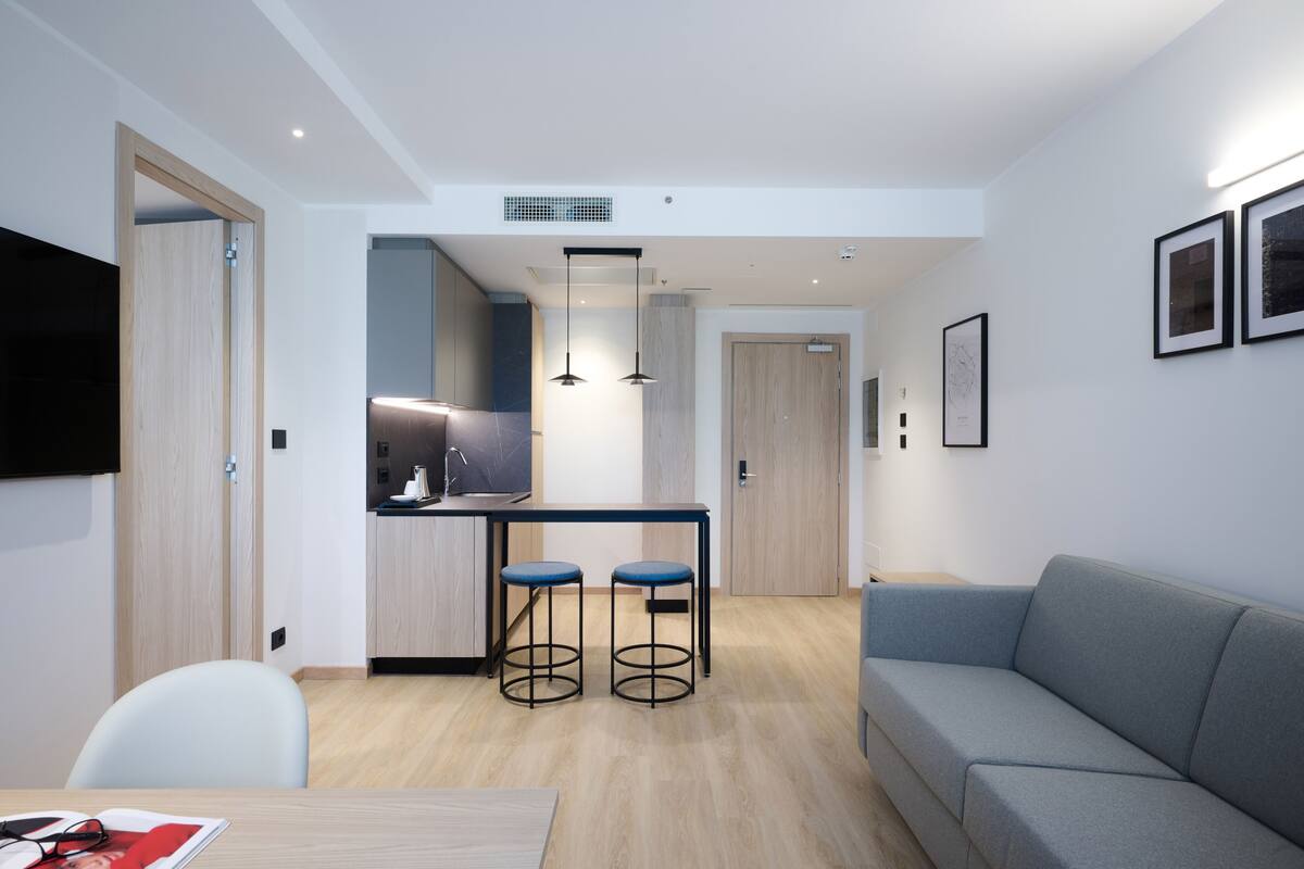 Una sala moderna con un sofá gris, una mesa de comedor de madera con dos taburetes azules, una zona de cocina con encimera negra y un televisor montado en la pared.