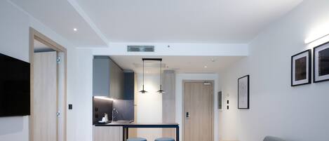 Una sala moderna con un sofá gris, una mesa de comedor de madera con dos taburetes azules, una zona de cocina con encimera negra y un televisor montado en la pared.