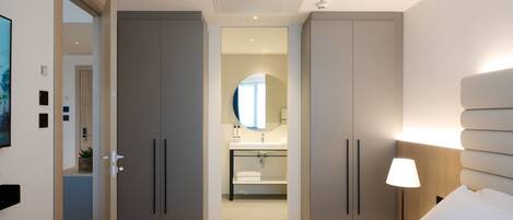 A modern hotel room with a large bed, a bedside table with a lamp, a wardrobe, and a bathroom visible through an open door.